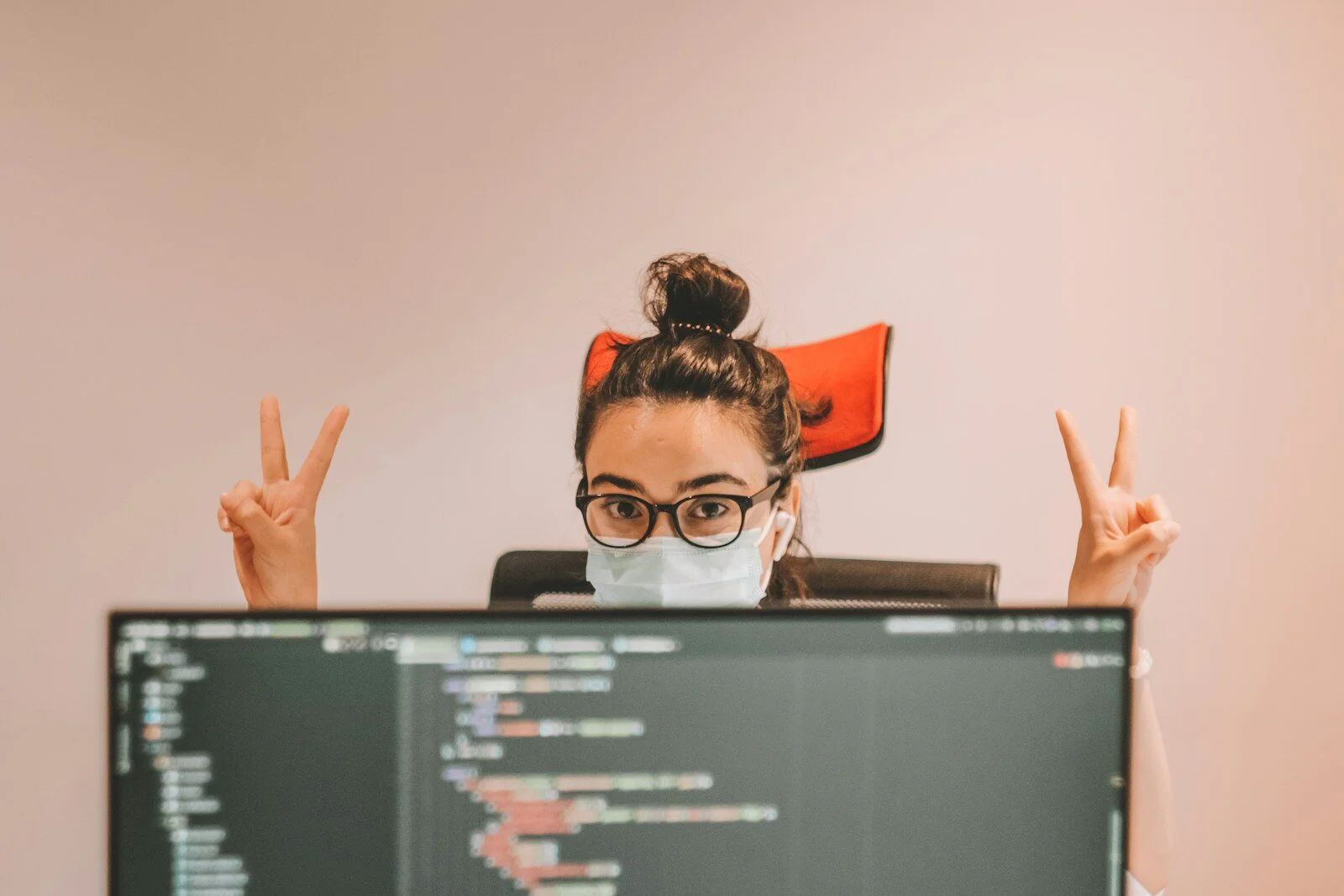Woman in black framed eyeglasses doing peace sign over monitors showing code